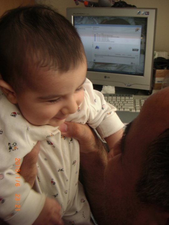 The image depicts a tender moment between a child and an adult. The child appears to be a baby, likely a little boy, given his attire which includes a white outfit with a pattern on the chest. The adult is holding the baby from the back, creating an intimate and nurturing scene. Behind them is an old-style computer screen showing a webpage, indicating this photo was taken in an indoor setting, possibly a home or an office. The date and time stamp at the top left corner of the image read "2/8/2012 23:27," suggesting the moment captured is from the past.