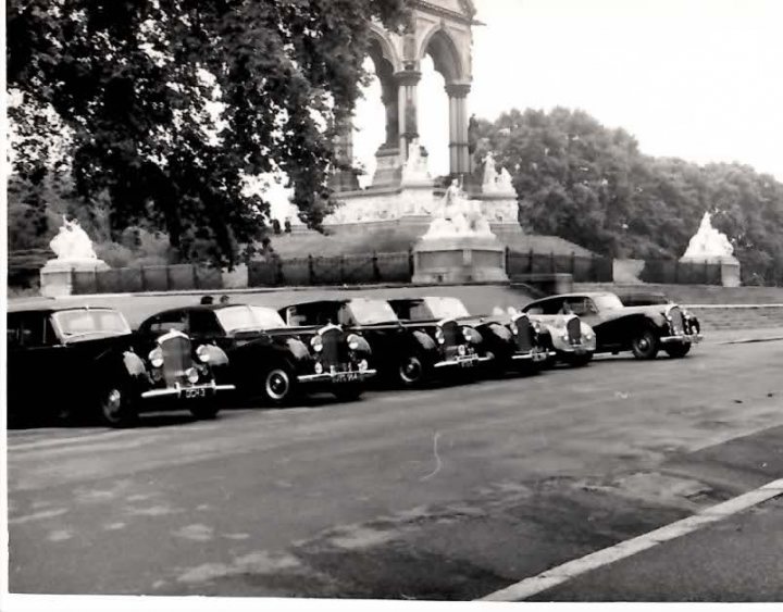 B&W Bentleys.... - Page 1 - Rolls Royce & Bentley - PistonHeads - The image is a black and white capture of a serene park scene featuring a row of vintage cars. The cars are neatly parked along a roadside, lined up in front of a prominent statue in the background. A distinctive fountain, adding to the monuments, is also visible in the distance on a grassy expanse, adorned with lush trees and a majestic building. The image exudes a vintage feel, reminiscent of a bygone era.