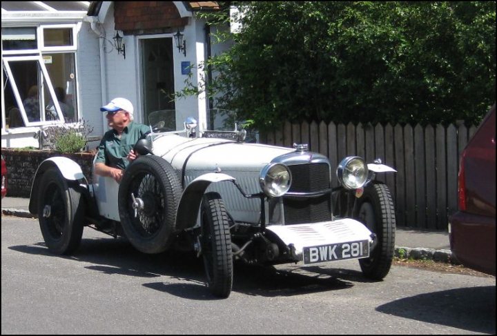 COOL CLASSIC CAR SPOTTERS POST!!! Vol 2 - Page 63 - Classic Cars and Yesterday's Heroes - PistonHeads - The image depicts a charming scene of a man driving a vintage white car down a street lined with traditional red brick houses. The car is a two-seater vintage model, characterized by its round tires and two large round lights for headlights. The man appears to be bald, wearing a blue cap and a green shirt, his attention directed forward as he maneuvers the old-fashioned vehicle. The license plate on the car reads 'BWK 281'. The background is filled with the quaintness of the residential area, with several folk-style houses and lush greenery adding to the idyllic atmosphere.