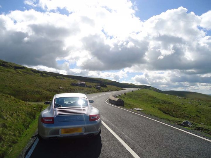 A car parked on the side of the road - Pistonheads - The image captures a serene landscape where a blue Porsche car is parked on the asphalt of a road. The car, positioned towards the left of the frame, is parked by the side of the road, which is bordering a lush green hillside. The road itself is marked with white lines and is flanked by a stone wall on the right side.

On either side of the paved road, there are grassy embankments that seem to be resting in the shade of the surrounding mountain peaks. These peaks, covered in green vegetation, provide a natural backdrop to the scene.

Above it all, the sky is filled with clouds, suggesting that the photo was taken during the day. Despite the presence of the car, the scene exudes a sense of tranquility and solitude. The car, the road, and the mountainous backdrop come together to create a harmonious composition that is pleasing to the eye.