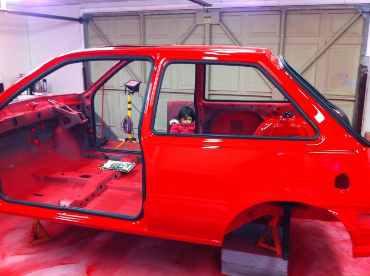 Fiesta RS Turbo Restoration - Page 7 - Ford - PistonHeads - The image depicts a vintage Toyota Land Cruiser from the 1960s being restored. The interior is a rich red color and the vehicle is at a car repair shop. A child wearing a red jacket is sitting in the passenger seat, adding a human element to the scene. The car's frame is visible outside, indicating that it is undergoing work on its chassis.