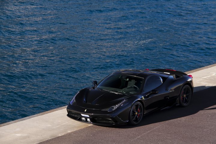 458 Speciale - Page 1 - Ferrari V8 - PistonHeads - This image is a dynamic photograph of a black Ferrari fastback, possibly a Lamborghini Adriatica Convertible, under a clear blue sky. The car is parked on the left side of a cement carport near the edge of a pier or jetty that extends over a body of water. The reflections of the vehicle suggest it's on a polished, smooth surface. The image captures the vehicle in beautiful natural light, highlighting its sleek and powerful design.