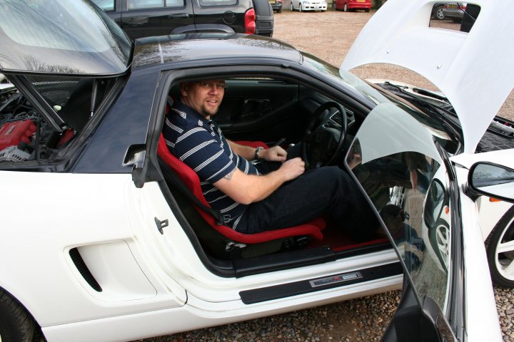 Pistonheads - The image depicts a man inside the driver's seat of a white convertible sports car with the top down, positioned to show a backward view. The man is casually dressed, with facial stubble, and either smiles or has a thoughtful expression. He holds the steering wheel, suggesting recent or imminent driving. The car is parked in a gravel area next to other parked vehicles, including cars and trucks, indicating a public space or parking lot.