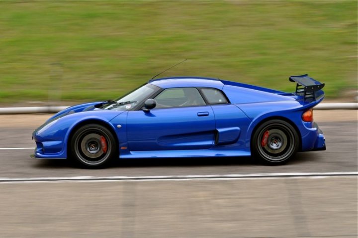 RE: Get Your Supercar Rides At Dunton Track Day - Page 5 - Events/Meetings/Travel - PistonHeads - The image features a sleek, blue sports car in motion on a paved road with grassy land portion visible to the side. The sports car is equipped with a prominent rear spoiler and a number badge on the side panel. The setting appears to be during daylight, as indicated by the bright and well-lit conditions. The car's design suggests speed and performance, typical of sports car models.