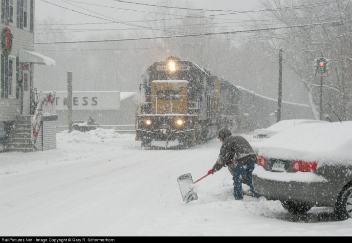 Tracks Ice Pistonheads - In the image, a man is valiantly shoveling snow from the road, perhaps attempting to clear a path for the train traveling behind him. The train's engine and the words "PRESS" are visible as it navigates the snow-covered tracks. A car is parked to the side, partially covered by the falling snow, adding to the wintry scene. The scene is set against a backdrop of houses nestled in the trees, which are blanketed in snow. The image captures the essence of a wintry day in a neighborhood affected by a snowstorm.