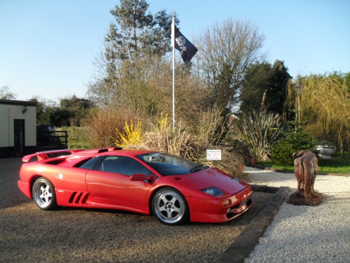 Who drives what? - Page 1 - East Anglia - PistonHeads - The image features a vibrant red sports car parked on a gravel driveway. The car, prominently occupying the left side of the image, is sleek and shiny, suggesting it's well-maintained. A large black flagpole stands behind the car, positioned centrally, while on the right side, a life-size bronze statue of a horse adds a unique touch to the scene. The setting appears to be a spacious, open area with a clear blue sky overhead and trees and bushes dotting the background, creating a sense of tranquility around the impressive vehicle.