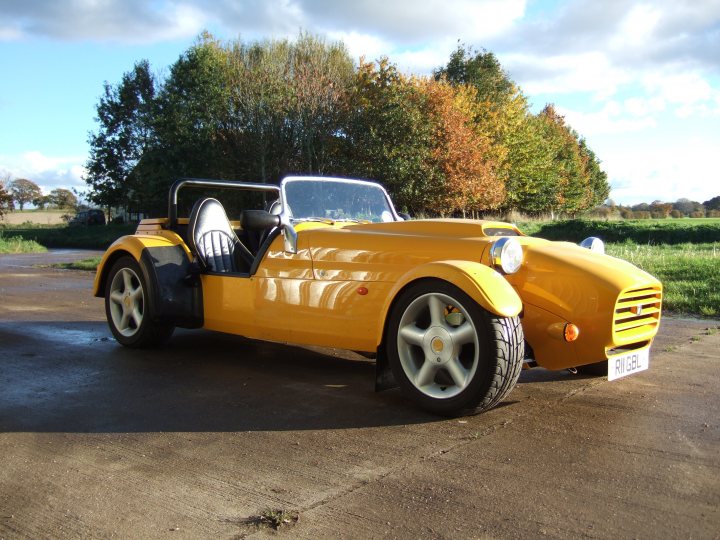 Pistonheads - This image features a distinctive yellow sports car parked on a concrete surface. The car's design has a classic yet modern feel, characterized by a smooth, curved body and a convertible top that appears to be up or folded back. The vehicle is sitting alone, with its wheels oriented to the side, suggesting it is stationary. The background is simple, with a grassy area hinting at a rural or semi-rural setting, and a line of trees with autumnal foliage providing a natural backdrop. The image has a high angle view, looking down at the car, which emphasizes its sleek lines and vibrant color.