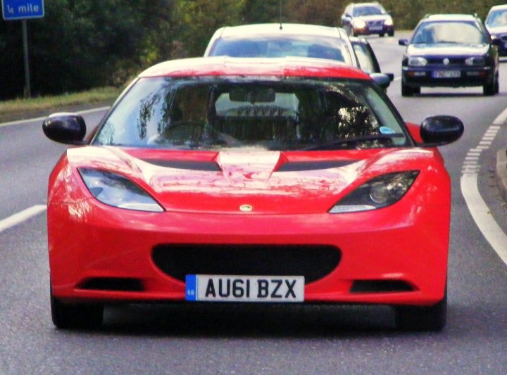 Pistonheads - The image shows a bustling scene on a wide highway, with two red sports cars being the focal point of the photograph. The sports cars, which appear to be Alfa Romeos, are moving towards the viewer, with the license plate AU61 BZZ clearly visible. The rest of the highway is filled with various types of other vehicles, indicating a busy traffic situation. The background features a clear sky and a few trees, suggesting that the highway is surrounded by greenery. The photograph has a blurred quality, especially visible in the upper portion of the image, perhaps due to the vehicle's speed and the photographer's focus on the near objects.