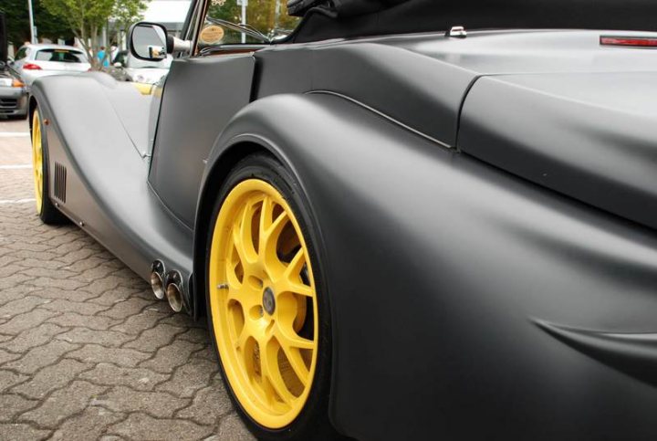 Service Pistonheads Sunday - The image features a dark, sleek sports car with a prominent yellow front tire that stands out against the vehicle's monochromatic exterior. The car appears to be at rest in a location with a sparsely used parking lot with other vehicles and trees in the background. The image captures a close-up view of the car's side profile, highlighting its aerodynamic design and the yellow tire's distinctive hue. The overall mood of the image is modern and fast-paced, embodying the allure of speed and luxury that such a vehicle represents.