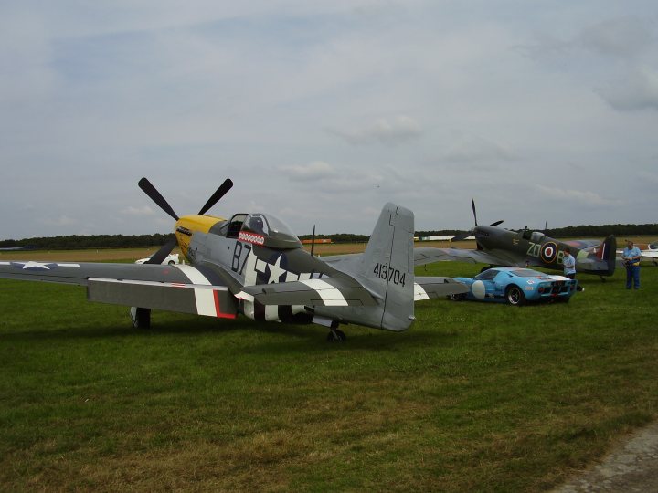 Day Pistonheads Asda - The image showcases a lively air show with two military-style planes prominently featured in the foreground. One plane has a sleek design with red and gray coloring, while the other sports a camouflage pattern and a yellow nose. They appear to be vintage or historic planes, as suggested by their craftsmanship and overall aesthetic. The planes are either on the ground or just taking off, and people can be seen gathering around them, enjoying the spectacle. The overall scene conveys a sense of excitement and nostalgia, characteristic of such outdoor aviation events.