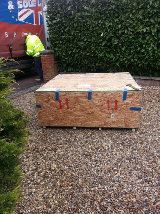 Delivery - Page 1 - Vipers - PistonHeads - The image shows a person, likely a construction worker, standing beside a freight truck, inspecting or possibly unloading a newly mounted platform. The platform in question appears to be a wooden structure with a green stripe along the top edge, and it is secured to a large cargo box. This box is placed outdoors on a gravel surface, with some shrubbery visible in the background. The freight truck from which the cargo box is being unloaded is partially visible in the left side of the image. The person's attention is focused on the cargo box, suggesting the situation may be an important or delicate task.