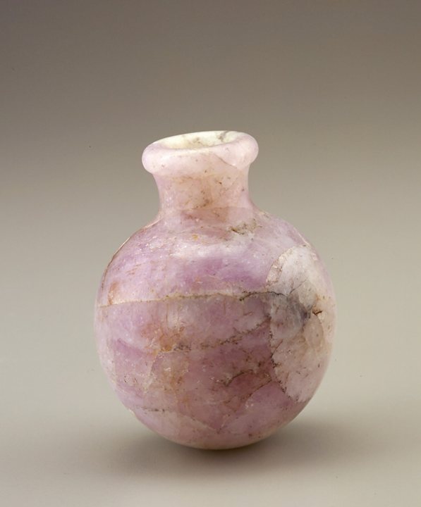 The image features a tall, cylindrical gray or pinkish vase with a visible opening at the top. The vase is situated against a neutral background, suggesting a focus on the object itself. There is a rough, cracked texture visible on the body of the vase, implying fragility or the effects of time. The style of the vase appears to be ancient, possibly suggesting a significant historical value or the inspiration from historical artifacts.