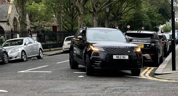 The image features a busy city street lined with parked cars on the right side of the road. The vehicles range in color and size, indicating a variety of makes and models. A prominent black Range Rover with a visible license plate is in the process of leaving its parking space, and appears to be pulling out of the parking line onto the road. There are no people visible in the frame, and the architecture and foliage suggest a dense urban environment. The condition of the road and the surroundings implies it is an active part of the street, likely a main thoroughfare.