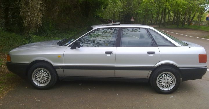 Pistonheads - The image shows a silver hatchback parked in a forested area with a road running alongside the parking area. The car is facing away from the viewer, with its side door open, suggesting a casual, possibly rural setting. The car features black wheels and a rear spoiler, typical of certain 80s or 90s era models. The surrounding foliage is thick and extends quite a bit into the background. The road and the edges of the parking area are visible, indicating that the car is properly parked in designated space. The setting appears peaceful and somewhat secluded, suggesting the photo may have been taken at a location removed from urban or densely populated areas.