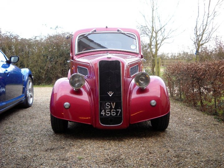 A red double decker bus parked in a field - Pistonheads - This image depicts a scene with two vintage cars parked on a driveway. The most prominent car in the foreground is a vintage red truck with a distinctive grille design. Its registration number "SV 4567" is visible on the grille. To the left of this car is another vehicle, partially visible and in a different color. The setting is outdoors with a clear sky and a lush green hedge in the background. The presence of the cars suggests it's a private property, with both vehicles parked on the gravel drive.