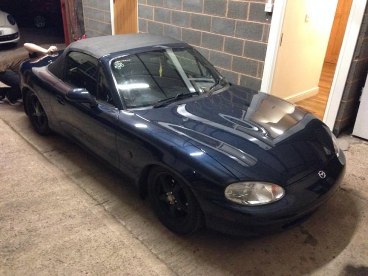 My MX5 - Its all abit of fun! - Page 1 - Readers' Cars - PistonHeads - The image shows a shiny, metallic blue Mazda MX5 Miata parked inside a garage or workshop. The car is positioned at a slight angle to the camera, giving a clear view of its side profile and the engine compartment. Its hood is open, and it appears to be in a state of disassembly or repair, with various tools and parts scattered around and a person working on it. The background consists of the garage wall and a doorway with an inner frame visible. The lighting in the room illuminates the car and indicates an indoor setting, contrary to the outdoor environment often suggested.