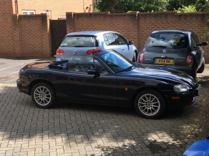 A car parked on the side of the road - Pistonheads - The image depicts an outdoor scene during the day. There are two cars parked side-by-side on a paved area. The first car, which is parked on the left side of the photo, is a black Volkswagen Beetle convertible. The second car, parked on the right side, is a silver hatchback with a distinctive grille and round headlights. This car appears to be a Ford. The cars are positioned in front of a brick wall, and behind them, there is a brick fence and a clear, blue sky. The style of the image is a straightforward, casual photograph without any artistic filters or dramatic angles.