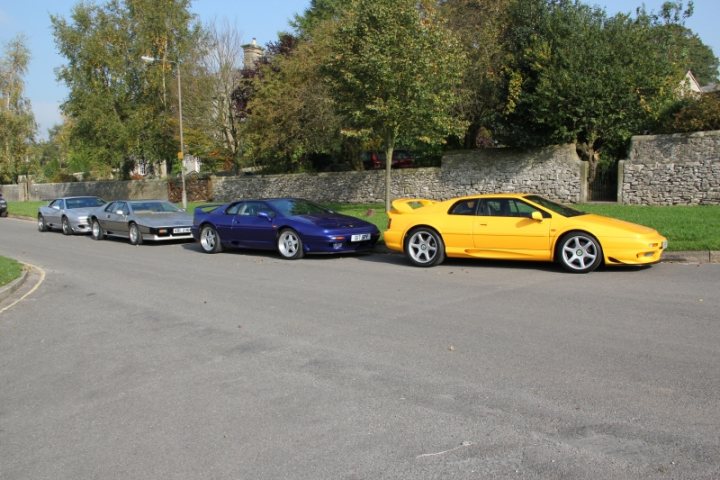 Esprit Pistonheads Bakewell - This image depicts an outdoor street scene featuring several cars parked alongside a sidewalk. The cars are a mix of colors and models, and they appear to be of similar vintage, possibly luxury or sports cars. The road is paved, and there's a grassy area and a stone wall visible in the background, suggesting a suburban or rural setting. The sky is partly cloudy, indicating it could be a middle-season day. The photo itself has a blueish hue, which may be due to the lighting conditions at the time the photo was taken.