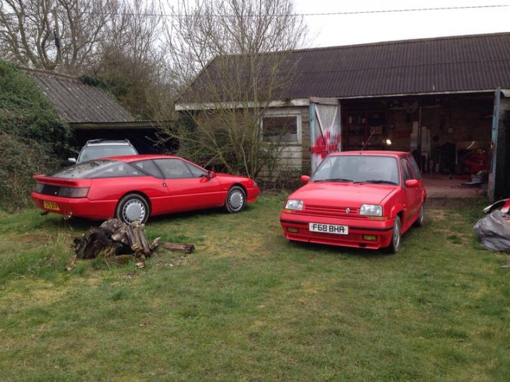 A red truck is parked in a field - Pistonheads - The image depicts two vintage cars parked on a grassy area next to a wooden building. The left car is a convertible in a shade of red, while the car on the right is painted a similar red but appears slightly older and smaller in size. The grass is green, and straw can also be seen scattered around. The wooden structure behind the cars has a connecting passage to another building. The overall scene is peaceful and appears to be a personal or residential area.