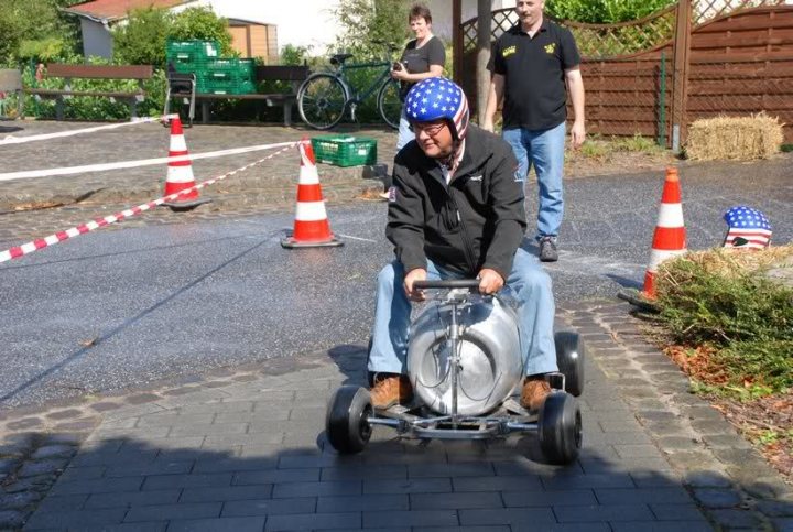 A man riding a motorcycle down a street - Pistonheads - In the image, a man is seated in a small go-cart, also known as a tricycle. He is wearing a blue helmet for safety and a black jacket for warmth. The go-cart is speeding down a street, which is lined with cones, indicating that he might be participating in a go-karting race or event. A young man, standing off to the side, appears to be watching the spectacle. The scene suggests a casual, leisurely atmosphere where the man in the go-cart is enjoying the ride.