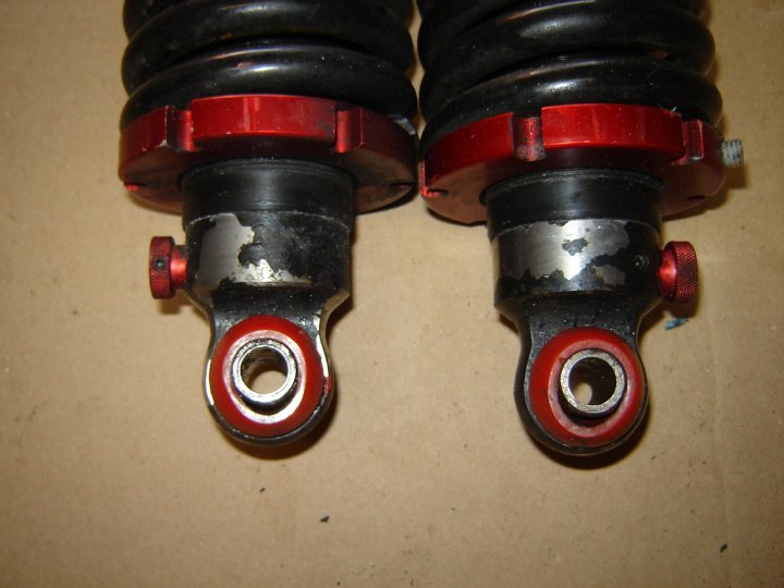 Question Avosprings Pistonheads - The image displays a pair of hydraulic dampers. They are shaped similarly to coils, with a main black body and a red base that includes the piston handle. The metal connector on the right damper is bent slightly and has some roughness on its surface, indicating wear or rough contact. The dampers appear ready for installation.