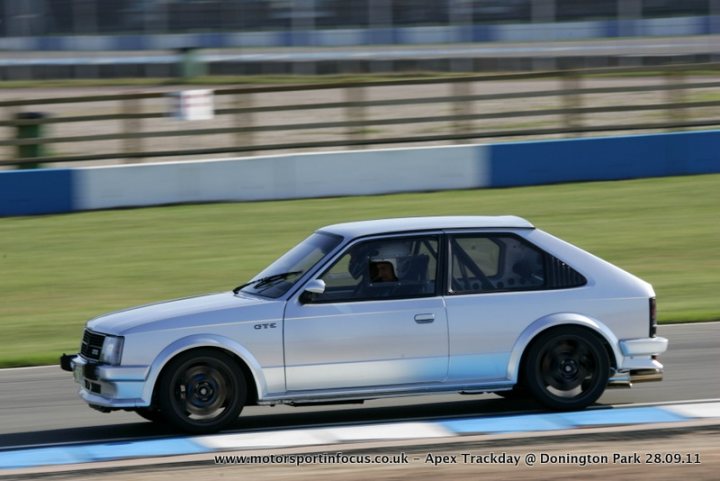 Pistonheads - The image features a silver two-door sports car. The car is in motion on a racetrack, depicted as driving around a corner with the windows open, suggesting a fast pace. The racing track is equipped with safety barriers and there's a blue edge to the track. The driving line appears to be marked by a white stripe, which is common in racing circuits.