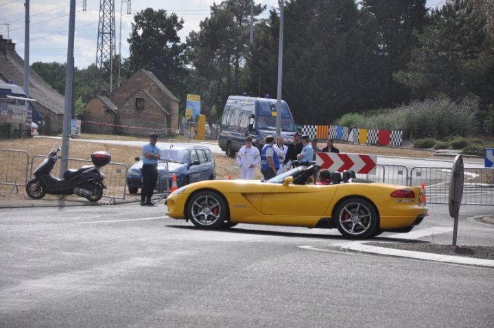 Le Mans 2011 - Page 1 - Vipers - PistonHeads - The image captures a vibrant street scene with a yellow sports car as the focal point. The car is stopped at a red light, marked with a red and white triangle. A group of people are standing on the side of the road, engaged in various activities. To the left of the image is a black moped. A few trees can be seen in the background, and there's a power pole nearby. The overall atmosphere suggests a typical day in an urban area.