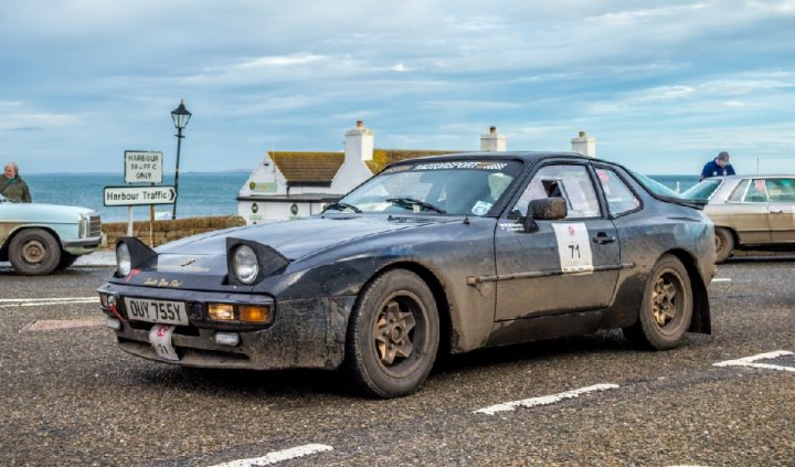 New 944 Arrival - Page 1 - Front Engined Porsches - PistonHeads - In the image, a vintage black car with round headlights is driving down a grey road. Behind the car, a beige station wagon is following. The road is somewhat worn with white lines marking separate lanes. On the left side of the image, there's a person standing, possibly waiting to cross. In the backdrop, you can see calming blue water and houses, suggesting a coastal town setting.