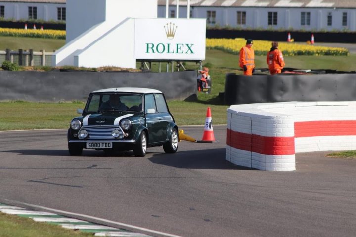 Pictures of your Classic in Action - Page 23 - Classic Cars and Yesterday's Heroes - PistonHeads UK - The image captures a dynamic scene on a race track. At the center, a green car is in motion, driving past a row of red and white barriers. A person wearing an orange jacket can be seen walking beside these barriers. In the background, there's a white building with a sign that reads "Rolex". The overall setting suggests a professional motorsport event, possibly a rally or circuit race.