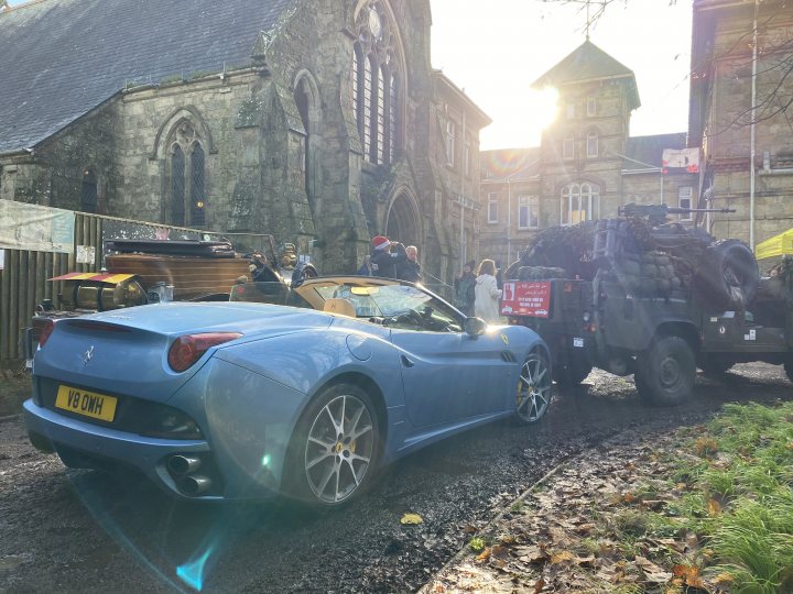 Ferrari California - Page 4 - Readers' Cars - PistonHeads UK - The image shows a vibrant blue sports car parked on the side of a street. It's an Aston Martin DB11, identifiable by its sleek design and distinctive grille. There are people nearby, suggesting they might be admiring the car or possibly attending an event. In the background, there is a church with a tall tower, and a truck can be seen further down the street. The setting appears to be in a small town or village, as indicated by the quaint architecture and the quiet atmosphere of the scene.