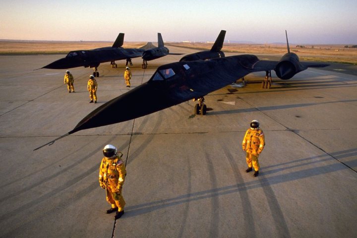 Post amazingly cool pictures of aircraft (Volume 2) - Page 422 - Boats, Planes & Trains - PistonHeads - The image shows a group of fighter jets parked on a tarmac. There are three main jets, with two additional aircraft visible in the background. The planes are painted in a dark color and have visible cockpits. In front of the aircraft, there is a group of individuals standing, dressed in protective suits and helmets that resemble astronaut gear. These figures appear to be inspecting or preparing the jets, given their attire. The environment suggests an airfield under clear skies during the daytime. There are no visible texts on the image.