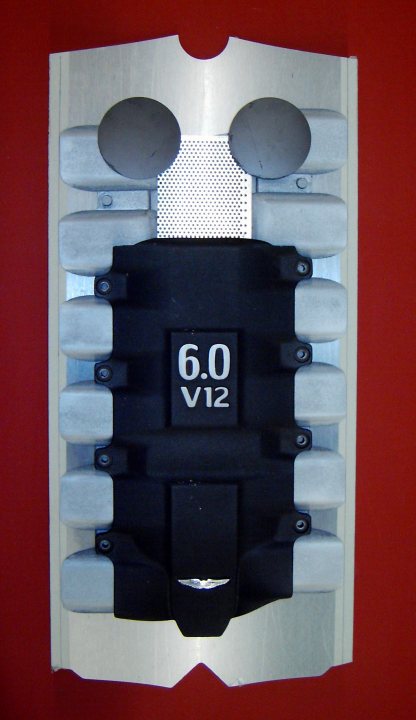 Martin Aston Pistonheads Clock Wall - The image shows a small, protective plastic container with multiple slots designed to hold screws or other small parts. Inside the container, there are various items, including what appears to be a black plastic piece with some markings or writing on it. The container is mounted on a red background, and the slots for holding screws are visible, suggesting it's a tool-related object used in activities requiring organization and easy access to small hardware. The specific content of the containers, other than the black piece, is not clearly distinguishable.