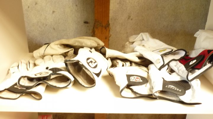 A pair of shoes and a pair of scissors - Pistonheads - This image shows a white shelf under a wooden panel, holding a collection of sports and protective gloves. The first gloves in the center are black and white, with a crescent-shaped design, and they are likely football gloves due to their size and shape. To the right, there is also a pair of red and white gloves, which are smaller and likely intended for boxing or martial arts. On the left side of the shelf, there is an open pair of white gloves, which appear to be designed for archery or similar sports. The overall style of the image is straightforward, providing no visible text and relying on the viewer to identify the types of gloves based on their shapes and colors.