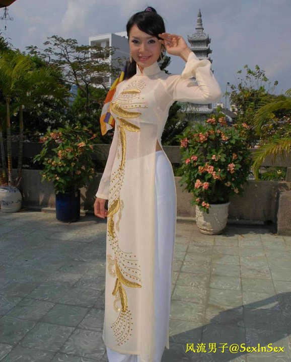 The image features a woman standing on a paved surface. She is wearing traditional Asian clothing, which consists of a long, white outfit adorned with gold trim. The woman has dark hair and is holding a small item in her hand, near her face. In the background, there is a well-manicured garden with flowering plants, and a stately building with Chinese architectural features. The setting appears to be a peaceful outdoor area, possibly in an urban park.