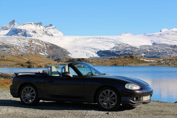 Respraying the Mk1 - what colour? - Page 1 - Mazda MX5/Eunos/Miata - PistonHeads - The image captures a serene outdoor scene. Dominating the foreground is a black convertible sports car, parked on the left with its top down, inviting the viewer to imagine an open-air drive. Behind the car, a calm lake stretches out, its surface reflecting the clear blue sky above. In the distance, snow-capped mountains rise majestically, adding a sense of grandeur to the landscape. The car's precise location on the side of a road by the water's edge suggests a journey paused to take in the beauty of nature.