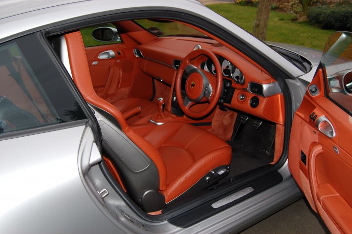 Opinion Pistonheads Terracotta - The image displays the interior of a car with the vehicle door open. The interior of the car features a two-door cabin with two red seats, one in each door side. The seats have orange insets, and the occupants side door rides low with two arm rests extended outward. A gearshift is visible partway inside the cabin, possibly indicating a manual transmission. The car's bodywork, which is painted in a shade of orange that includes a brighter tone on the side, is partially visible as it transitions to another color on the hood and roof.