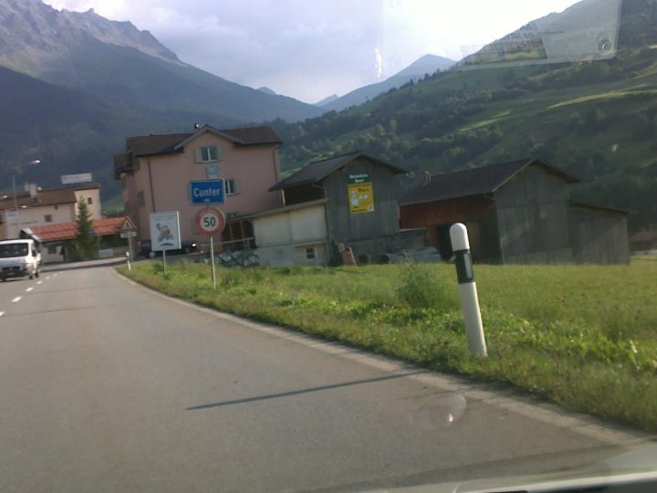 Pistonheads Towns Swiss Amusinglynamed - The image captures a rural street in an alpine region. Dominating the scene is a large, pink-colored house with a red roof, tucked between the surrounding mountains and a grassy field. The house is located neatly on the side of the road, which is flanked by another building and topped with a speed limit sign. The landscape is predominantly covered in lush greenery, and the presence of a few vehicles and traffic signs indicate a minimal amount of traffic. The angles of the buildings and the perspective of the photograph make it seem as if the house is being swallowed up by the verdant, mountainous terrain.