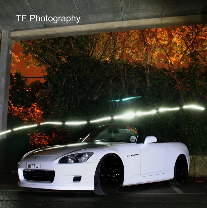 time we had pictures of everyones jap wagons - Page 80 - Jap Chat - PistonHeads - The image depicts a white sports car parked in a concrete area under a bridge. The lighting appears to be artificial or natural nightlight, as it creates a dramatic effect with the car's headlights illuminating the space. The car is positioned at an angle to the camera, emphasizing its design features such as the front grille and wheel arches. In the background, there are trees and foliage, indicating an outdoor parking area. The image is watermarked with "TF Photography", suggesting it was taken by a photography service called "TF Photography."