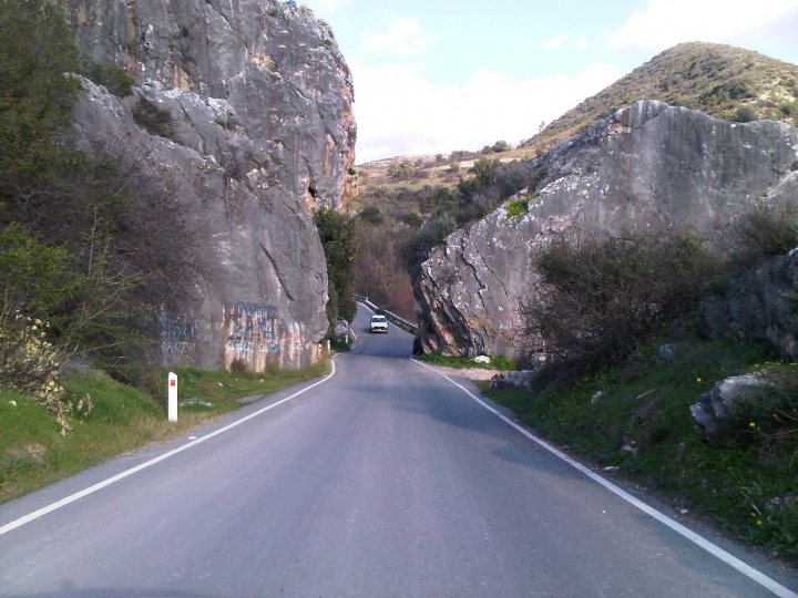 Hello from Cyprus! - Page 1 - Aston Martin - PistonHeads - The image provides a first-person view of driving down a narrow, winding road surrounded by majestic, craggy cliffs. The road seems to be in the middle of nowhere and either leads into a mountain or overlooks a valley. The sky is partly cloudy, suggesting it might be a cool day. There's no traffic, adding a feeling of tranquility and solitude to the scene.