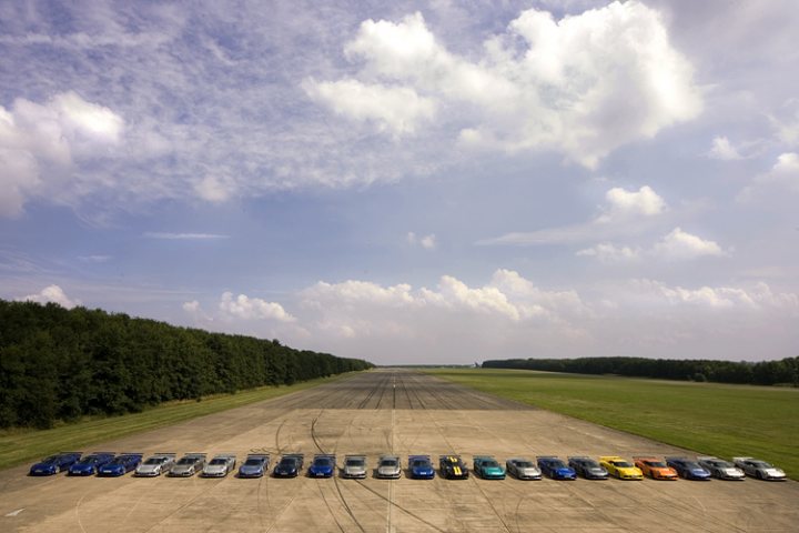 Chose Noble Pistonheads - The image shows a collection of cars lined up in a straight line, facing the camera, on a runway with a backdrop of clouds and trees. It appears to be a photograph rather than a painted composition. The street appears empty and the lighting suggests either early morning or late afternoon sunlight, the different cars have different colors and designs, suggesting they could be part of a show or rally.