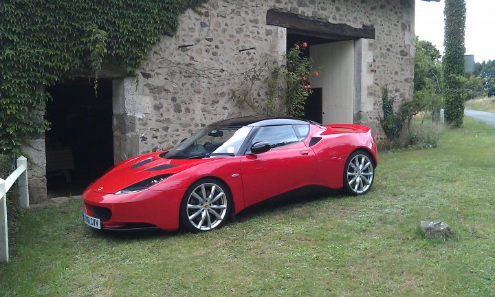 Dave the Trimmer does the Evora - Page 1 - Evora - PistonHeads - The image shows a red sports car parked next to a building. The car is sleek and has the roof down, indicating a convertible. The car's wheels are visible and they appear to be standard, not the large, high-performance variety that one might expect on a sports car. The building behind the car has a distinctive appearance with large stones and ivy growing up its side. There's an open doorway or archway at the base of the building, suggesting the vehicle is parked quite close to the entrance. The setting is outdoors on grass, implying a peaceful, non-urban environment.