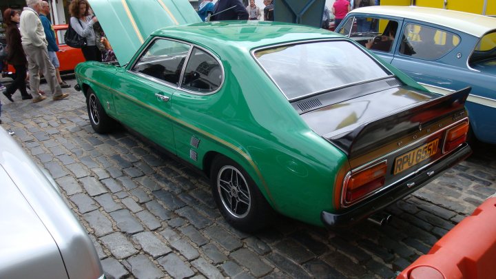 Classic Pistonheads Faversham - The image displays a vibrant vintage car show, where a variety of classic and hot rod cars are on display on a cobblestone street. The bright lighting emphasizes the shiny surfaces and chrome details of these vehicles. In the foreground, a pristine green hot rod with a sleek, forceful design and a prominent rear spoiler is the centerpiece. Its lower stance indicates it might be a race car or heavily modified, turning attention towards its unique appearance. The atmosphere is lively, with people standing near the cars, likely admiring their design and history. The scene is predominantly outdoors, with a brick pavement providing a contrasting texture to the reflective surfaces of the cars.