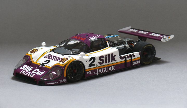 Tamiya 1/20th Jaguar XJR9-LM - Page 14 - Scale Models - PistonHeads - In the image, a sleek silver and black race car is the main focus. The car is adorned with a vibrant purple stripe running along its body. The logo "2 Silk Jaguar" is prominently displayed on the car, indicating its association with Jaguar's racing team. Above the car, there's a clear blue sky with a fluffy white cloud, creating a serene backdrop for the car's vibrant colors. The setting appears to be an outdoor museum or exhibition, given the open sky and the replica nature of the car on display.