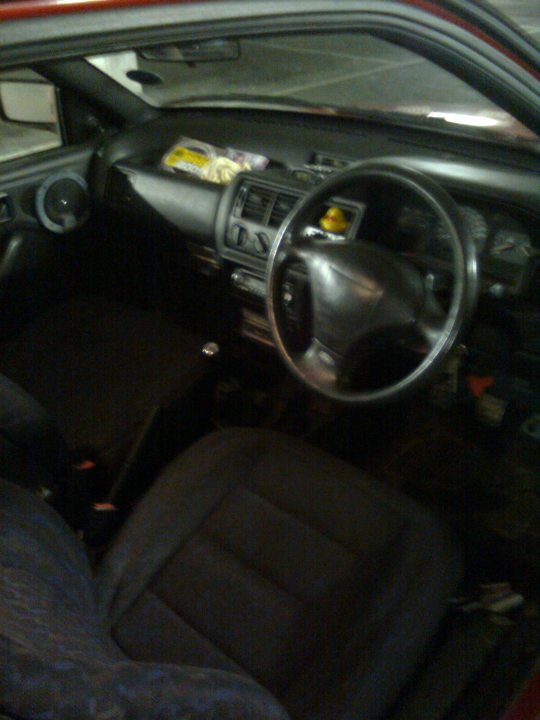 Pistonheads Escort - The image shows an interior view of a vehicle. The driver's seat is on the left side of the shot, indicating a car likely made for countries where drivers drive on the left side of the road. The seat appears to be made of a fabric material and is pushed up to allow the driver to enter. A control panel, possibly a stereo or navigation system, is mounted in front of the steering wheel, suggesting the vehicle has a variety of onboard entertainment and guidance systems. The dashboard features gauges and indicators typical for a vehicle, and a glove compartment is visible as well. The car's interior has a dark, perhaps black or very dark gray, color scheme.