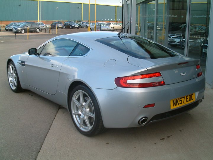 Hello All - Page 1 - Aston Martin - PistonHeads - The image depicts a silver Aston Martin DBS parked on the pavement in front of a building. The car has a sleek design with a dark roofline and distinctive taillights. It is equipped with a pair of rearview mirrors and silver alloy wheels. The license plate is clearly visible and reads "NK57 EDC". The car is surrounded by other vehicles parked in the vicinity, indicating that this could be a parking lot or a similar public space.