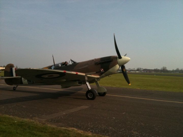 Spitfire Pistonheads Suspension - This image captures a moment of an orange and green Bell P-39 aircraft, specifically a commander's pilotless trainer aircraft, resting on a runway. The airplane features a brown camouflage pattern and has a propeller with two blades visible at the front. It is parked on concrete, and there is a figure near the aircraft, likely an aircraft enthusiast or aviation professional. The runway lies on a clear day with a patch of green grass visible in the background, adding a vibrant contrast to the mechanical subject of the image.