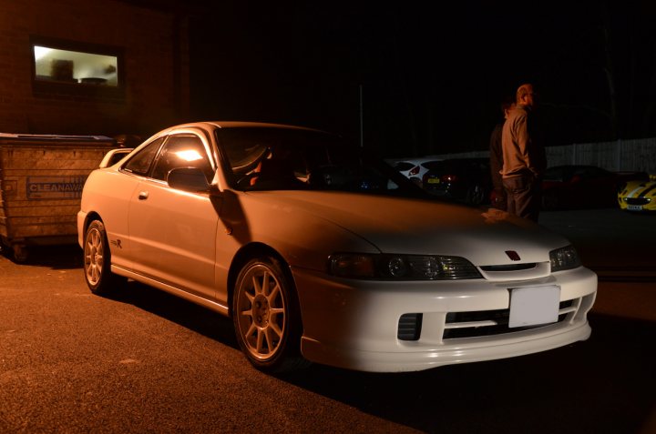 Pistonheads March Essex - The image presents a tranquil night scene featuring a silver sedan parked on a street. The car has a clean and polished exterior, possibly a hatchback or a smaller vehicle. A person is standing by the car, partially visible, suggesting a gathering or an event taking place nearby. The surrounding environment is dimly lit, mainly from streetlights, and there's a fence with a dumpster visible in the background, indicating an urban or semi-urban setting. The person and the car cast long shadows on the ground, indicating the presence of artificial lighting, possibly from street panels or vehicle headlights.