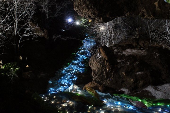 Santa in a cave  - Page 1 - South West - PistonHeads - This image showcases an enchanting underground stream, surrounded by towering, ice-covered rocks. A long line of blue lights, resembling tiny stars or glowing orbs, leads the eye towards a hidden light source beyond the frame. This fantastical scene is further enhanced by the lush greenery above-ground and the warm illumination from the light bulbs embedded in the rocks, casting an ethereal glow against the stark gray and white landscape. The scene is both miniature and majestic, inviting the viewer to imagine exploring such an otherworldly wonderland.