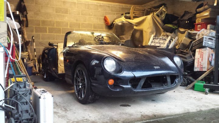 A car is parked next to a parking meter - Pistonheads - The image shows an indoor storage area, possibly a garage or a spare room, filled with various items. The main subject is an old, black car that dominates the center of the frame. The car is parked in front of a brick wall, and there's clutter all around, creating a sense of disarray. In the background, there are glimpses of other objects, including what might be a bicycle and various boxes or containers. The room appears to be quite crowded, with limited space between the items and the car.