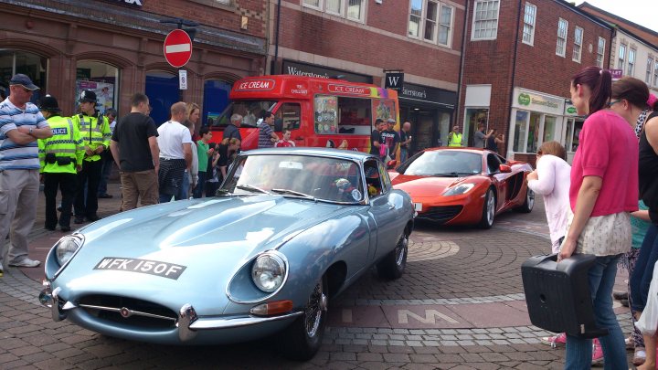 Ormskirk MotorFest - Page 1 - North West - PistonHeads - The image captures a lively street scene in what appears to be a city center. A vintage sports car and a red vehicle are the main attractions, parked on a roundabout. Several people are standing nearby, engaged in conversation, creating a sense of community and shared interest. A large red truck can be seen in the background, adding to the urban bustle. The juxtaposition of the vintage cars and the red truck suggests a blend of old and new in the cityscape.